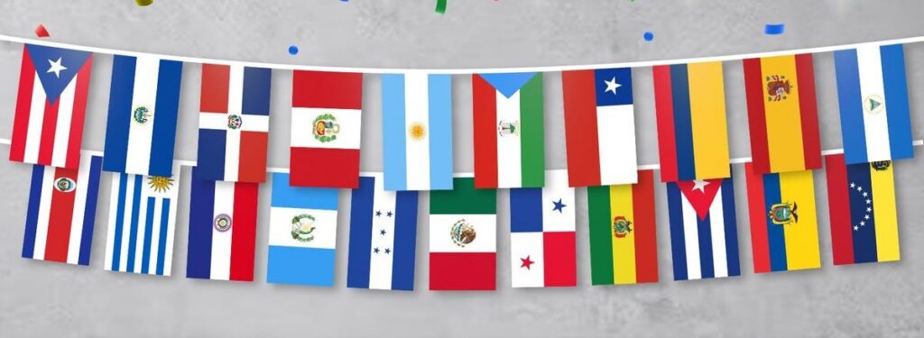Una guirnalda festiva de aproximadamente 20 banderas nacionales, predominantemente de países hispanohablantes de América Latina y España, cuelga en dos filas superpuestas sobre una cuerda blanca. Las banderas son coloridas y presentan los diseños distintivos de cada nación. Entre ellas se observan la bandera de Chile, Argentina, Perú, Colombia, México y España. El foconfetindo es una pared de textura gris clara. Pequeños trozos de confeti de colores brillantes —rojo, verde, azul y amarillo— caen dispersos por la parte superior de la imagen, añadiendo un toque de celebración.