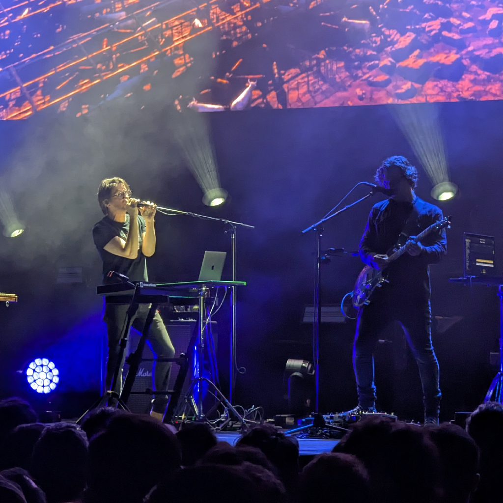 Una escena de concierto en vivo muestra a dos músicos en el escenario bajo luces brillantes. A la izquierda, un hombre con gafas y camiseta oscura canta con las manos sobre el micrófono y está de pie detrás de un soporte con un teclado y un portátil. A la derecha, otro hombre con pelo rizado y ropa oscura toca una guitarra eléctrica, con un micrófono delante. El escenario está oscuro, iluminado por focos blancos y azules. Detrás de los músicos, una gran pantalla proyecta una imagen abstracta en tonos anaranjados y marrones, que parecen estructuras o fragmentos. Las cabezas del público son visibles en silueta en la parte inferior.