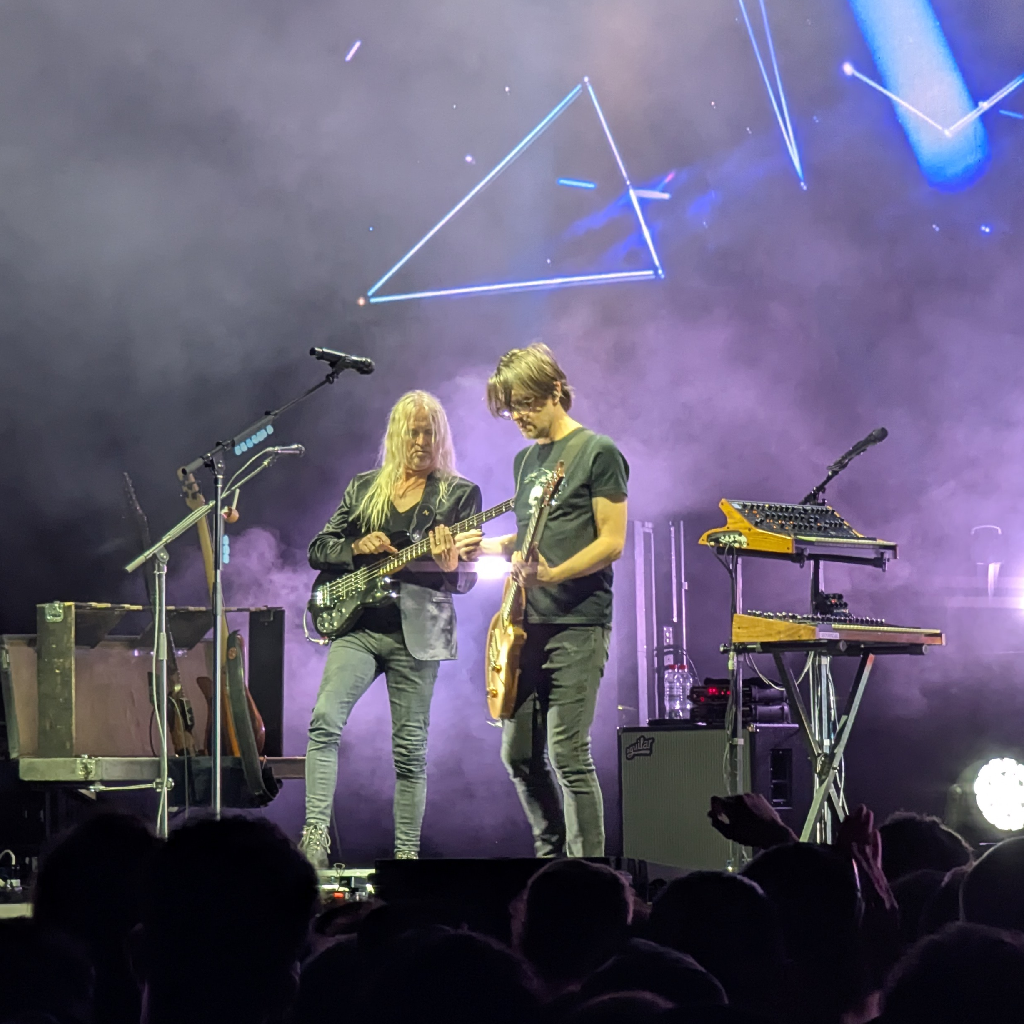 Una escena de concierto en vivo con dos músicos en el escenario, iluminado con luces moradas y azules. En el fondo brumoso, se proyecta el contorno brillante de un triángulo blanco y otras líneas geométricas.
A la izquierda, un hombre de cabello largo y rubio platino, vestido con una chaqueta de cuero negra y jeans oscuros, toca un bajo eléctrico negro. A su lado derecho, otro hombre con cabello castaño de longitud media y gafas, vistiendo una camiseta oscura y jeans, toca una guitarra eléctrica de color claro. Ambos músicos están concentrados en sus instrumentos.
A la derecha del segundo músico, hay un soporte con dos teclados o sintetizadores. En la parte inferior de la imagen, se ven las siluetas de la audiencia con algunas manos levantadas, indicando un público animado.