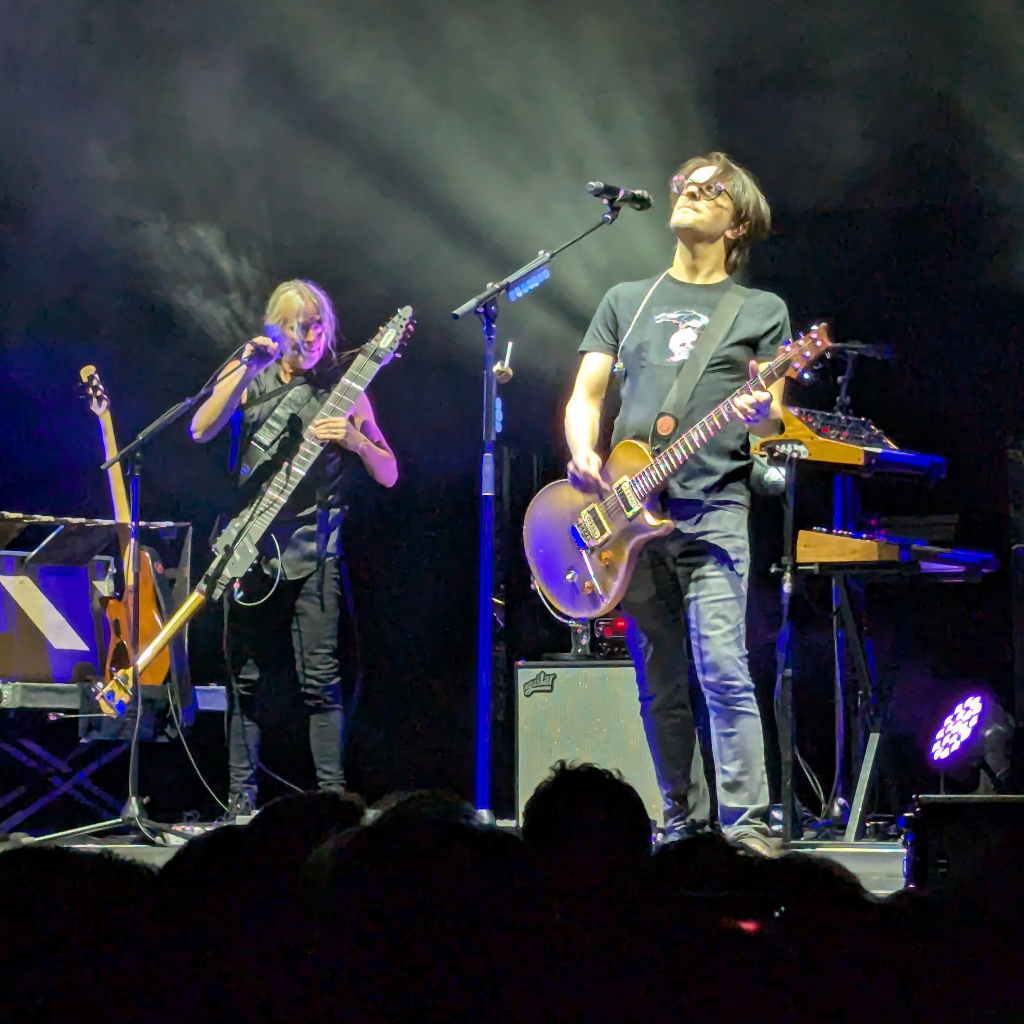 Una escena de concierto en vivo con dos músicos principales en el escenario, ambos bajo focos de luz blanca en un fondo oscuro y con humo.
A la derecha, un hombre de cabello oscuro y gafas de sol, vistiendo una camiseta negra con un diseño sutil y jeans oscuros, toca una guitarra eléctrica de color dorado o bronce. Mira hacia arriba, con la cabeza ligeramente inclinada.
A la izquierda, otro músico de pelo rubio y corto, con una camiseta sin mangas oscura y pantalones negros, toca un instrumento de cuerda largo y oscuro, similar a un bajo o un Chapman Stick, mientras canta en un micrófono.
En el fondo se aprecian soportes de micrófono, un amplificador blanco y teclados electrónicos. La parte inferior de la imagen muestra las siluetas de la audiencia.