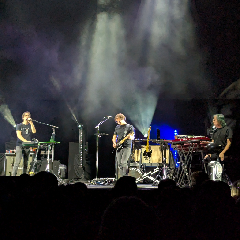 Una imagen de un concierto de rock en vivo, tomada desde la perspectiva de la multitud, que muestra a una banda de tres músicos en un escenario oscuro pero bien iluminado.
A la izquierda, un baterista con camiseta oscura toca un gran kit de batería de color cobre y plata. En el centro, un bajista de pelo largo y rubio, vestido con un chaleco negro y jeans oscuros, toca un bajo eléctrico; cerca de él, se ven varias guitarras acústicas apoyadas. A la derecha, el cantante principal y guitarrista, con pelo castaño medio y gafas, lleva una camiseta negra con un diseño claro y toca una guitarra eléctrica de color claro mientras canta en un micrófono de pie. A su lado, hay un teclado en un soporte.
El escenario está iluminado con potentes focos que proyectan haces de luz a través de una ligera neblina o humo, creando una atmósfera dramática. En primer plano, las siluetas oscuras de la multitud se ven con algunas manos alzadas, indicando que están disfrutando del espectáculo.