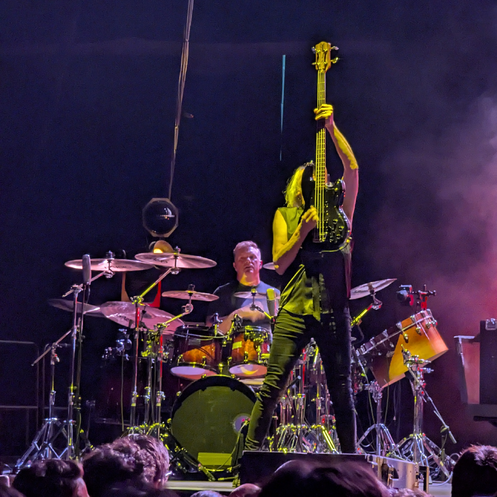 Una imagen de un concierto en vivo con dos músicos en el escenario. En primer plano, a la derecha, un bajista de pelo largo y claro levanta su bajo eléctrico negro verticalmente sobre su cabeza con ambas manos, ocultando su rostro. Viste ropa oscura. Detrás y a la izquierda, un baterista de mediana edad, con pelo corto, está sentado tocando un gran set de batería con múltiples platillos y tambores. Mira hacia adelante, concentrado. El escenario está oscuro, pero los músicos están iluminados por luces de concierto que crean un ambiente vibrante, con una luz rojiza/morada difusa en el lado derecho. Se aprecian las cabezas del público en la parte inferior de la imagen.